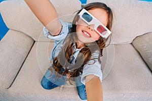 Stylish brightful selfie portrait of joyful little girl in 3d glasses with long brunette hair smiling to camera on couch