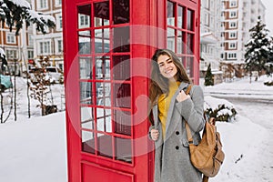 Stylish british portrait of charming young woman with long brunette hair walking on street near red telephone box on
