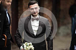 Stylish brunette smiling groom in a suit with a brown bow holds