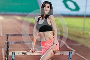 Stylish brunette woman in pink shorts and tank top posing for the camera on running track near the barriers running jumping at the