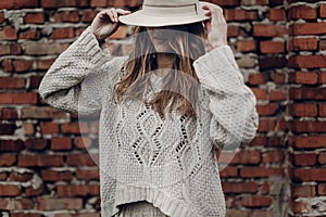 Stylish brunette woman in white hat and boho white sweater posing near red brick wall