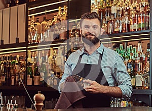Stylish brutal barman is cleaning the glass with a cloth at bar counter background.