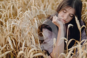 Stylish calm woman sitting in wheat field in evening light. Atmospheric tranquil moment. Young female in rustic linen dress