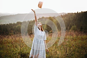 Stylish carefree boho girl throwing her hat in the sky in sunny light  at atmospheric sunset. Happy hipster woman in linen rustic