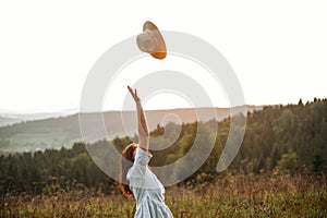 Stylish carefree boho girl throwing her hat in the sky in sunny light  at atmospheric sunset. Happy hipster woman in linen rustic