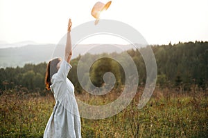 Stylish carefree boho girl throwing her hat in the sky in sunny light  at atmospheric sunset. Happy hipster woman in linen rustic