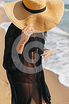 Stylish carefree woman holding hat and relaxing on sunny beach at sea, close up. Summer vacation. Young fashionable female in