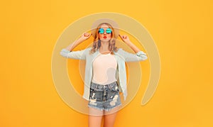 Stylish caucasian blonde young woman posing wearing straw hat, shorts and shirt on yellow background