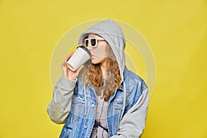 Stylish Caucasian girl in jeans and glasses 8 bit on a yellow background. Holding a disposable coffee cup to go and drinks. Copy