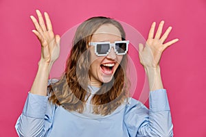 Stylish caucasian girl in a pale blue t-shirt and 8-bit glasses waving arms to the top on a pink background