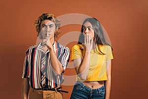 Stylish caucasian man with a hushing gesture and a young surprised female against a brown background