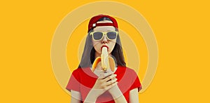 Stylish caucasian young woman eating banana wearing red baseball cap on orange background