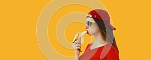 Stylish caucasian young woman eating banana wearing red baseball cap on orange background