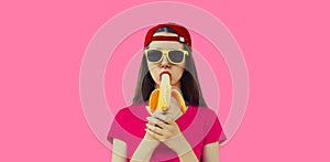 Stylish caucasian young woman eating banana wearing red baseball cap on pink background