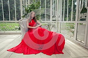 Stylish celebrity woman in red silky dress sitting in white summer pavilion