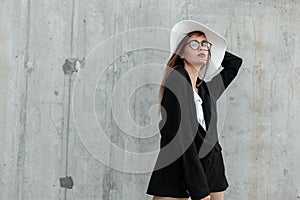 Stylish charming young woman in vintage glasses in fashion black-white clothes and straw hat posing on gray background in street.