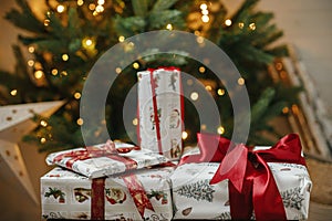 Stylish christmas gifts at christmas tree with golden lights bokeh. Wrapped christmas presents with red ribbon under decorated
