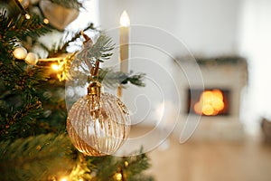Stylish christmas gold bauble on tree close up against burning fireplace. Beautiful decorated christmas tree with vintage