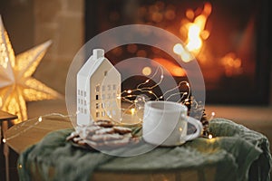 Stylish christmas little house with golden lights, gingerbread cookies and tea cup on table on background of fireplace in festive