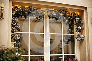 Stylish christmas mistletoe branches with golden  festive lights on window store at holiday market in city street. Christmas