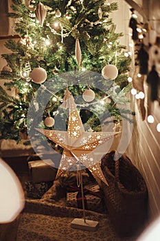 Stylish Christmas star, tree with white baubles, boho ornaments, golden lights and gifts in atmospheric evening room. Festive