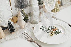 Stylish Christmas table setting. Cedar branch with bell on plate, vintage cutlery, wineglass, modern christmas trees and houses on