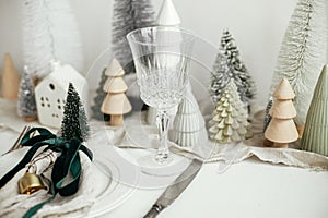 Stylish Christmas table setting. Festive napkin with ribbon and bell on plate, vintage cutlery, wineglass, modern christmas trees