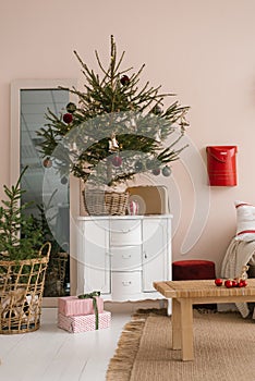 Stylish Christmas tree with toys in a wicker basket stands on a white chest of drawers in the interior of the living room