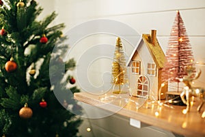 Stylish christmas wooden house, glitter christmas tree and golden reindeer in festive lights. Miniature fairy village, modern
