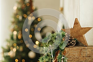 Stylish christmas wooden star, spruce branches on fireplace mantel close up on background of christmas tree lights. Festive rustic