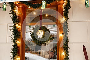 Stylish christmas wreath with fir branches and lights at front window of store at holiday market in city street. Space for text.