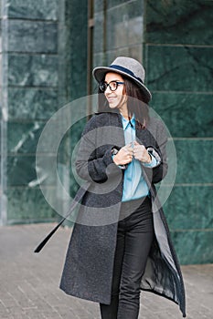 Stylish city portrait fashionable young woman walking in long grey coat on street. Wearing hat, black glasses, smiling
