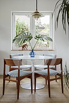 Stylish composition of dining room interior with design table, chairs, tropical leaf in vase, window and elegant decoration.