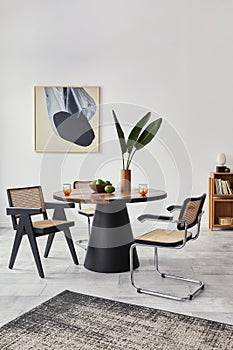 Stylish composition of dining room interior with design table, modern chairs, decoration, tropical leaf in vase, fruits, bookcase.