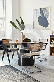 Stylish composition of dining room interior with design table, modern chairs, decoration, tropical leaf in vase, fruits, bookcase.