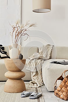 Stylish composition at fancy interior with wooden coffee table, dried flowers in vase, slippers, basket and book in modern home.