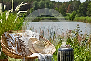 Stylish composition of outdoor garden on the lake with design rattan armchair, coffee table, plaid, pillows, drink. Elegant.