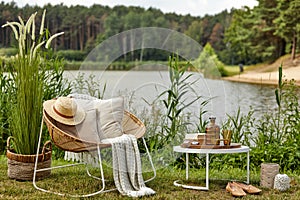 Stylish composition of outdoor garden on the lake with design rattan armchair, coffee table, plaid, pillows, drinks and elegant ac