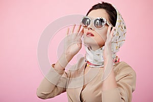 Stylish confident brunette woman touching his sunglasses, wearing in scarves, looking up, isolated on a pink background. photo