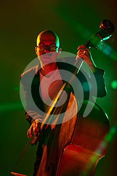 Stylish, contemporary bassist in glasses performs in red-green neon stage light against gradient studio background.