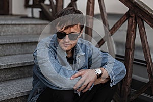 Stylish cool young man hipster in blue denim clothes in trendy sunglasses sits on a vintage staircase in the city