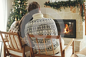 Stylish couple in cozy sweaters relaxing at fireplace with festive mantle on background of stylish decorated christmas tree with