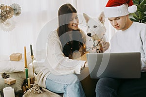 Stylish couple with cute white dog in lights and santa hat celebrating christmas holidays at home