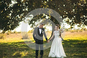 Stylish couple of happy newlyweds walking in field on their wedding day with bouquet. In the middle of the field ther is