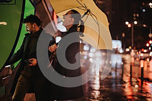 Stylish couple having a conversation on a rainy day while one person withdraws cash from an ATM machine using an