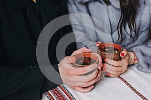 Stylish couple holding cups with hot tea in hands on wooden porch in winter snowy mountains. Happy romantic family with drinks. H