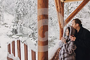 Stylish couple holding hot tea in cups and looking at winter snowy mountains from wooden porch. Happy romantic family with drinks