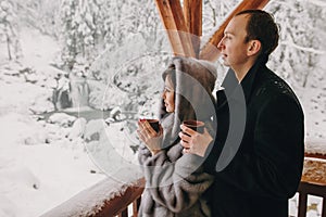 Stylish couple holding hot tea in cups and looking at winter snowy mountains from wooden porch. Happy romantic family with drinks