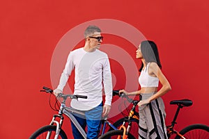 Stylish couple of man and woman with bicycles stand on the background of a red wall and look at each other in love, wearing