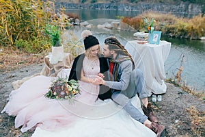 Stylish couple newlyweds are relaxing on a plaid and sitting before a lake. Bride and groom with dreadlocks are posing